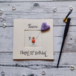 Handcrafted personalised 50th birthday card for her, female pebble figure holding a wine glass and a purple heart balloon. Created on cream card.