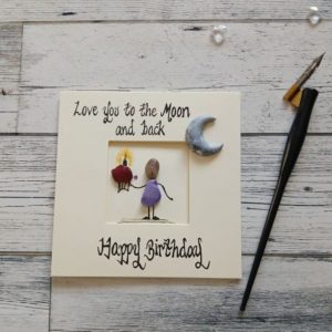 Happy Birthday card for the one you love, handmade pebble art card, Love you to the moon and back, Happy Birthday. Female pebble figure holding a cake and placed in the corner of the card a handpainted moon shaped pebble. Created on cream card.
