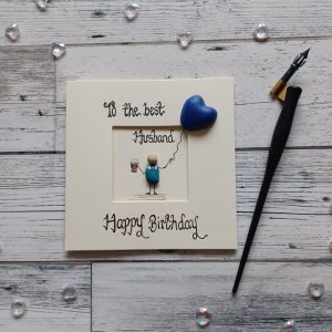 Birthday card for husband, handwritten in black ink, to the best husband Happy Birthday, handmade pebble art birthday card. Male pebble figure holding a beer and a blue heart shape balloon. Created on cream card.
