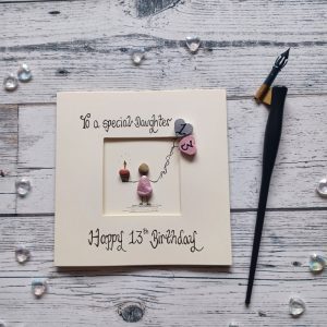 13th birthday card for a special daughter, pebble art card,pebble figure girl holding a cake and 2 heart balloons. Handwritten in black ink, To a special daughter, Happy 13th Birthday. Created on cream card.