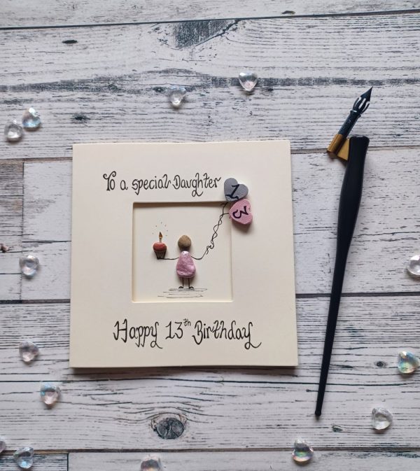 13th birthday card for a special daughter, pebble art card,pebble figure girl holding a cake and 2 heart balloons. Handwritten in black ink, To a special daughter, Happy 13th Birthday. Created on cream card.