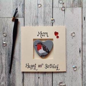 80th birthday keepsake card for Mum. Placed in the middle of the card is a handpainted heart shaped concrete pebble with a robin painted onto it. Created onto cream card.