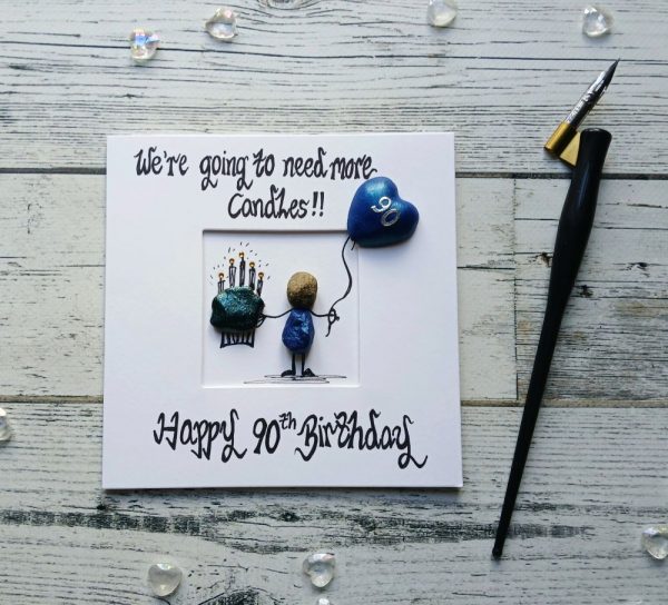 90th birthday card for him, a perfect card for a grandad or dad. Handmade pebble art card. Handwritten using black ink, We're going need more, Happy 90th birthday. Male pebble figure holding a big cake a blue heart balloon. Created on white card.