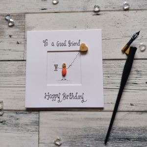 A birthday card for a friend, handmade pebble art card for female friend. Handwritten using black ink, To a good friend Happy Birthday. Female pebble figure holding a glass of wine and gold heart shape balloon. Created on white card.