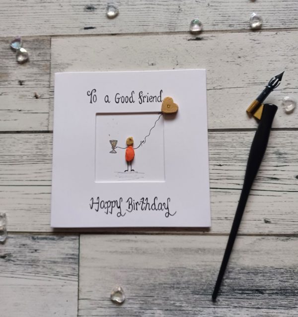 A birthday card for a friend, handmade pebble art card for female friend. Handwritten using black ink, To a good friend Happy Birthday. Female pebble figure holding a glass of wine and gold heart shape balloon. Created on white card.