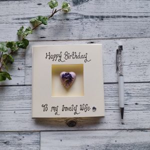 Happy Birthday card for your wife. Square cream triple-fold card, placed in the middle of the card is a pink heart-shaped concrete pebble decorated with a small bunch of lilac paper flowers. Handwritten the words happy Birthday to my lovely wife.