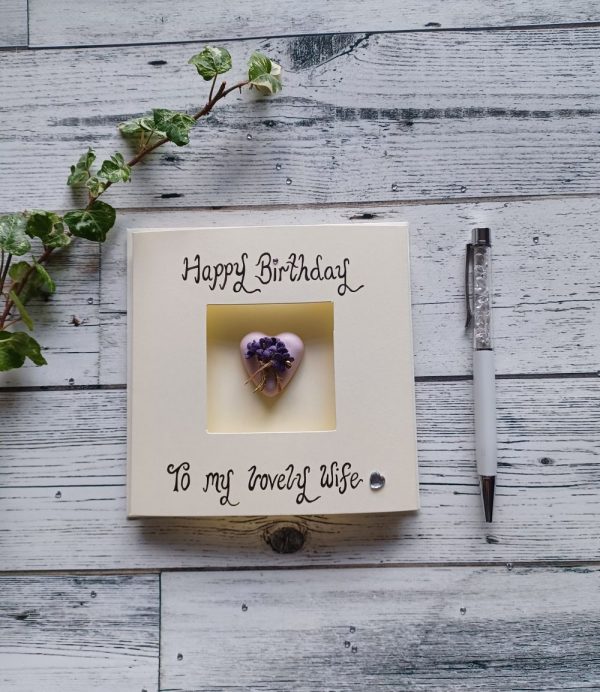 Happy Birthday card for your wife. Square cream triple-fold card, placed in the middle of the card is a pink heart-shaped concrete pebble decorated with a small bunch of lilac paper flowers. Handwritten the words happy Birthday to my lovely wife.