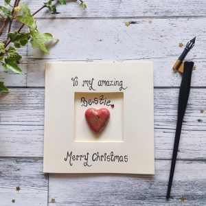 Handmade Christmas card for best friend, handwritten using black ink, To my amazing bestie, Merry Christmas. Placed in the centre of the card red heart shaped pebble. Created on cream card.