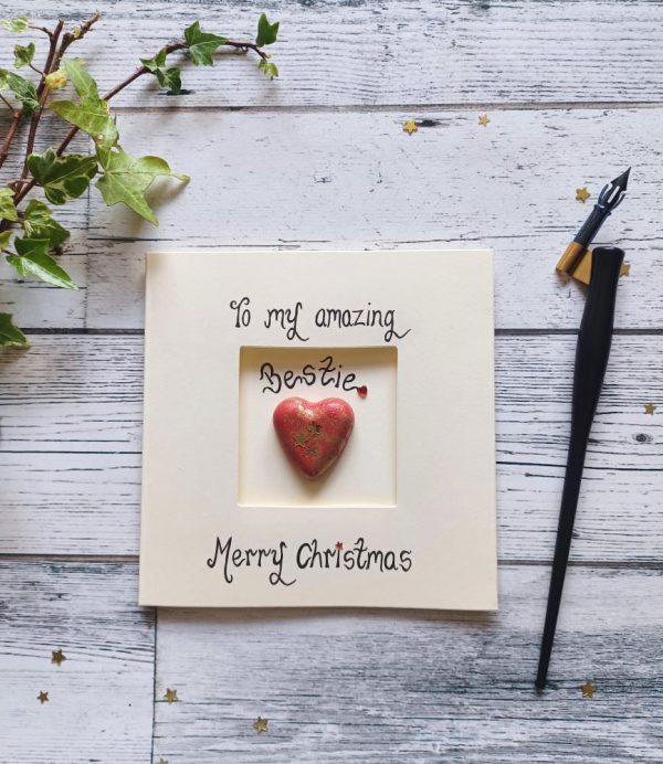Handmade Christmas card for best friend, handwritten using black ink, To my amazing bestie, Merry Christmas. Placed in the centre of the card red heart shaped pebble. Created on cream card.
