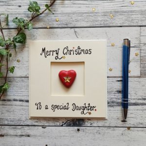 Handmade Christmas card for daughter, Handwritten using black ink, Merry Christmas, To a special Daughter. Placed in the centre of the card a red heart shaped pebble. Created on cream card.