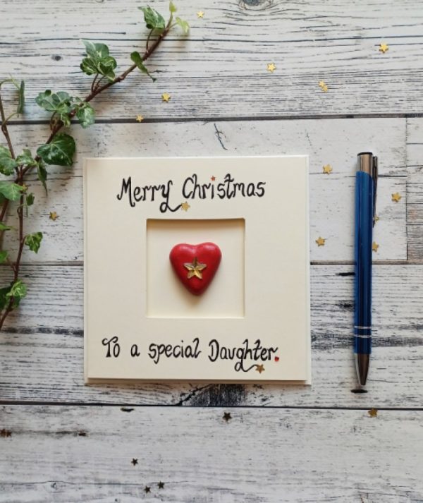 Handmade Christmas card for daughter, Handwritten using black ink, Merry Christmas, To a special Daughter. Placed in the centre of the card a red heart shaped pebble. Created on cream card.