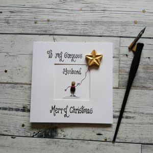 Handmade Christmas card for husband. Handwritten using black ink, To my gorgeous husband, Merry Christmas. Male pebble figure holding a gold star balloon. Created on white card.
