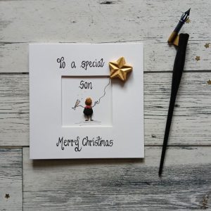 Handmade Christmas card for Son. Handwritten using black ink, To a special Son, Merry Christmas. Male pebble figure holding a gold star balloon and a glass of champagne Created on white card.