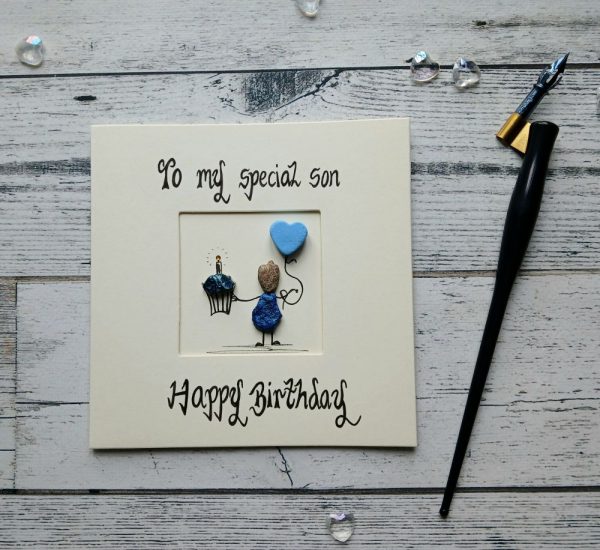 Handmade birthday card for son, handwritten using black ink, To my special Son, Happy Birthday. Male pebble figure holding a cake and a blue heart shape balloon. Created on cream card.